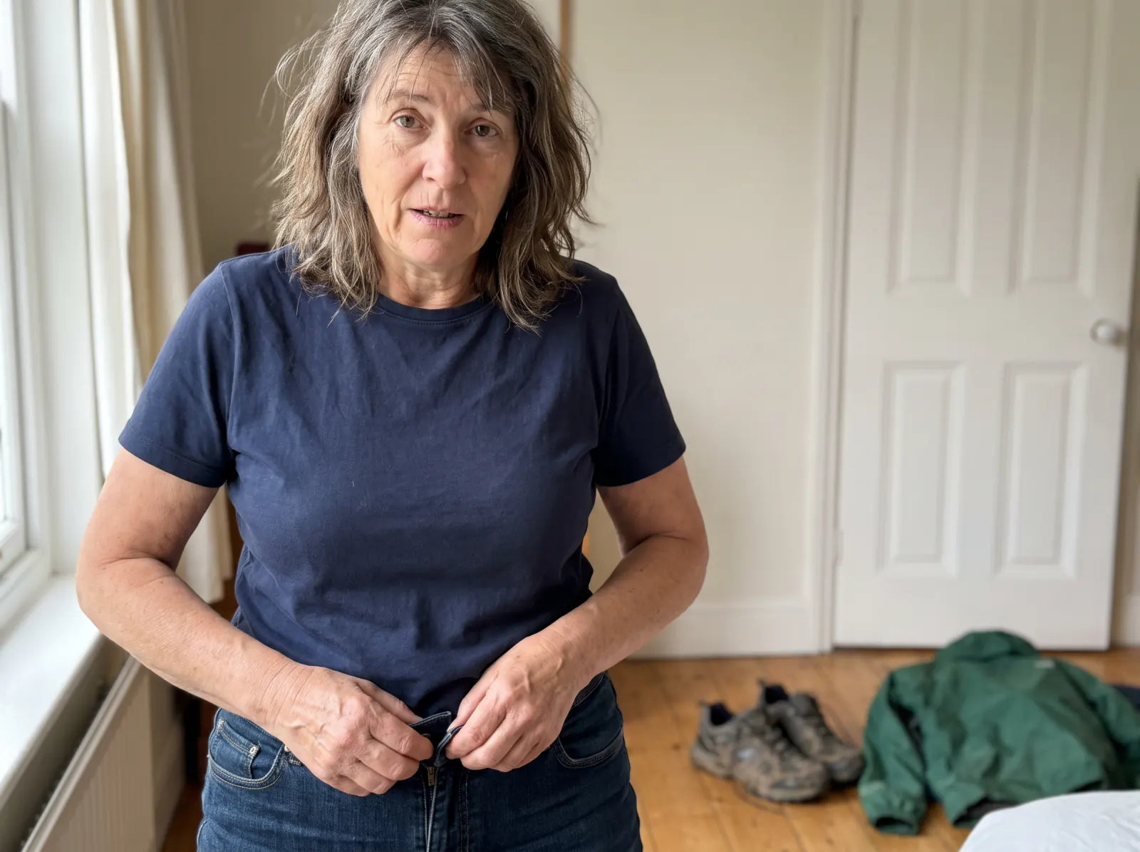 A woman over 40 fastening familiar jeans that now feel tight, with her walking shoes and jacket nearby.
